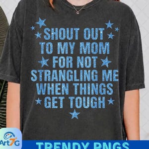 May include: A charcoal grey short-sleeve t-shirt featuring the blue glittery text SHOUT OUT TO MY MOM FOR NOT STRANGLING ME WHEN THINGS GET TOUGH, surrounded by blue glittery stars. The shirt has a crew neckline and a relaxed, vintage-washed finish.