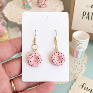 May include: A pair of dangle earrings featuring miniature pink doughnut charms with blue and red sprinkles. The doughnuts are attached to gold-toned metal fishhook wires with small jump rings. The earrings are displayed on a white rectangular card.