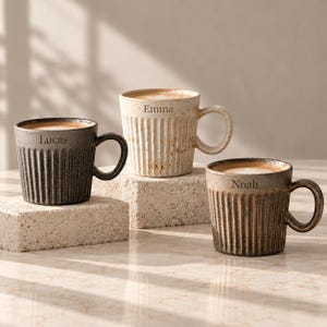 May include: Three ceramic mugs with vertical ribbed textures and rounded handles. The dark grey mug is engraved with the name Lucas, the cream-coloured mug with Emma, and the bronze-toned mug with Noah. Each mug contains a beverage with foam.