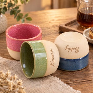 May include: Three ceramic cups with a matte beige base and glazed upper sections in pink, green, and blue. The green cup features the name Kenna engraved on the side, while the blue cup features the name Bobby engraved on the side.