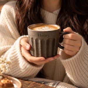 Puede incluir: Una taza de cerámica de color marrón oscuro con un exterior texturizado y acanalado, y un asa. El nombre Henry está grabado en la parte frontal de la taza, que contiene una bebida de café con un diseño de arte latte blanco.