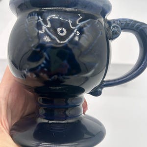 May include: A dark blue ceramic mug with a glossy finish, featuring a rounded bowl-shaped body, a thick curved handle, and a pedestal base. The glaze shows subtle variations in color intensity, creating a deep navy appearance with lighter highlights.