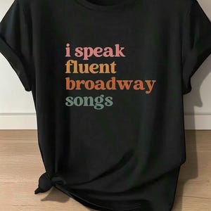 May include: A black short-sleeved t-shirt with a crew neckline and rolled sleeves. The front features the text "i speak fluent broadway songs" printed in stacked lines with "i speak" in pink, "fluent" in orange, "broadway" in rust, and "songs" in teal.