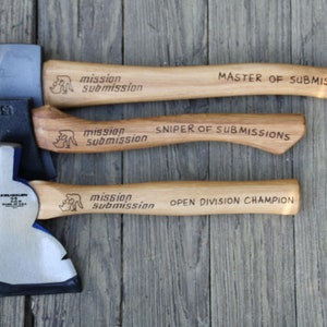 May include: Three axes with wooden handles and metal blades are displayed on a wooden surface. The handles are engraved with the words "mission submission" and titles such as "MASTER OF SUBMISSIONS".