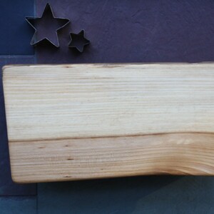 May include: A light wood cutting board with a natural edge. The board is rectangular and has a smooth surface. It is sitting on a blue and red tiled surface.