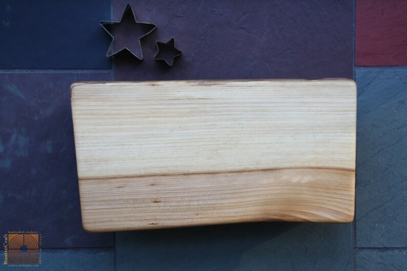 May include: A light wood cutting board with a natural edge. The board is rectangular and has a smooth surface. It is sitting on a blue and red tiled surface.