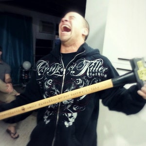 May include: A person holding a black and brown sledgehammer with a white and black graphic print on the handle. The graphic print says "Only Warmonger of the Bartow Fair". The person is wearing a black hooded sweatshirt with a white graphic print that says "Candy Killer Clothing".