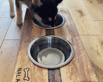 May include: A wooden dog bowl stand with two stainless steel bowls. The stand is stained with a dark brown wood stain and has a light brown wood inlay. The stand is engraved with the name "Nala" and a bone symbol.