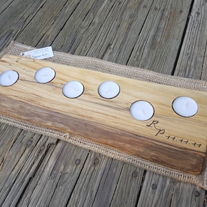 May include: A wooden candle holder with six cutouts for tealight candles. The holder is made of natural wood with a rustic finish and has the initials "R.P." and the date "11.11.11" engraved on the surface. The holder is sitting on a burlap fabric.