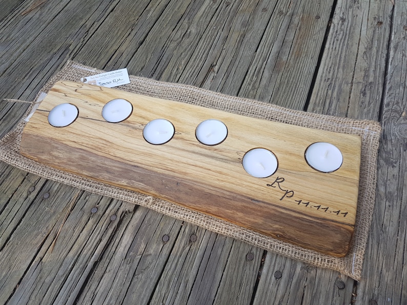 May include: A wooden candle holder with six cutouts for tealight candles. The holder is made of natural wood with a rustic finish and has the initials "R.P." and the date "11.11.11" engraved on the surface. The holder is sitting on a burlap fabric.