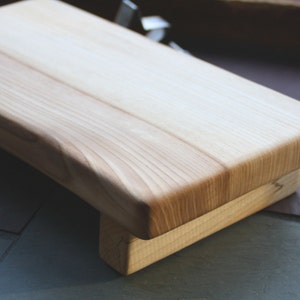 May include: A natural wood cutting board with a raised edge and a base. The board is made of light wood with a smooth finish.