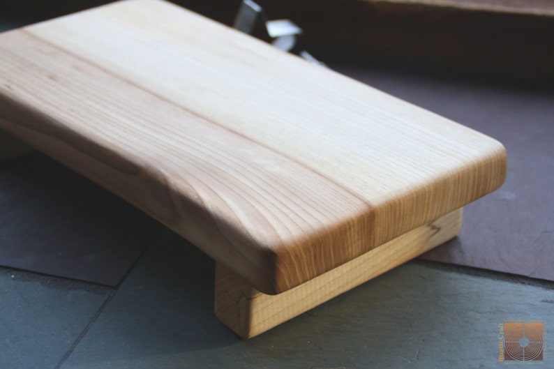 May include: A natural wood cutting board with a raised edge and a base. The board is made of light wood with a smooth finish.