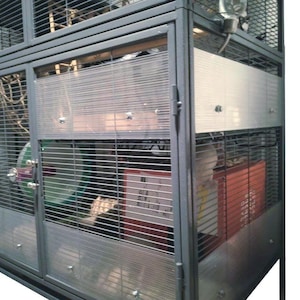 May include: A tall, dark gray metal animal cage featuring a wire mesh structure, a front-opening door, and solid panels. Inside, a green exercise wheel is visible alongside a red rectangular enclosure and a clear water bottle attached to the top.