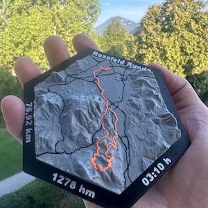 May include: A hexagonal 3D printed topographic map of the Rossfeld Runde trail. The grey terrain features black road lines and a bright orange path. Text includes Rossfeld Runde, 76.92 km, 1278 hm, and 03:10 h printed on the black border.