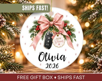 May include: A round white ceramic ornament featuring a pink ribbon bow, green pine branches, red berries, a black car key fob, and a scalloped tag with the text New Driver. The name Olivia and the year 2026 are printed in black script and serif font.