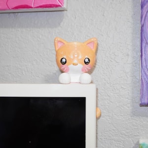 Peut inclure: Une petite figurine de chat en céramique orange et blanche avec de grands yeux noirs, des joues roses et des pattes blanches. Le chat est posé sur le bord supérieur d'un cadre rectangulaire blanc, avec une finition brillante et arrondie.