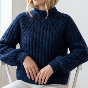 Könnte beinhalten: Ein dunkelblauer Langarmpullover mit vertikalem Rippstrickmuster, hohem Stehkragen und Raglanärmeln. Das Kleidungsstück hat eine dicke, strukturierte Strickkonstruktion mit gerippten Bündchen an den Ärmeln und einem gerippten Saum an der Taille.