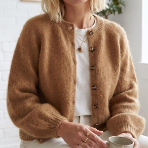 May include: A tan, long-sleeved, knitted cardigan featuring a round neckline and a front button closure with dark brown buttons. The cardigan is worn over a plain white t-shirt, and a gold-toned necklace with a small pendant is visible at the neck.