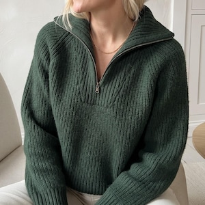 May include: A dark forest green long-sleeve sweater featuring a ribbed knit texture, a high collar, and a silver-toned metal zipper that extends from the neckline to the upper chest. The garment has long sleeves with ribbed cuffs and a straight hem.