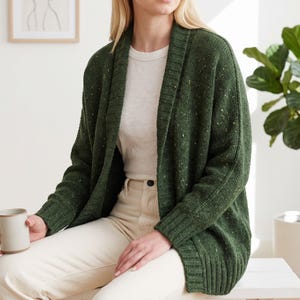 May include: A forest green open-front cardigan featuring a textured knit pattern with subtle multicolored flecks throughout. The garment has a wide ribbed shawl collar, long sleeves with ribbed cuffs, and a straight hemline with a ribbed finish.