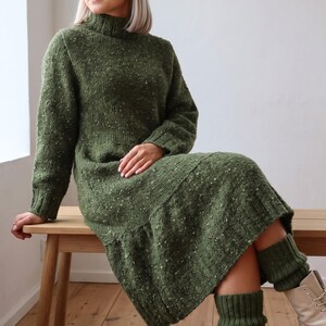 May include: A long-sleeved, olive green knit dress featuring a mock turtleneck, a textured speckled yarn finish, and a drop-waist silhouette. The garment includes matching ribbed knit leg warmers that extend to the mid-calf area.