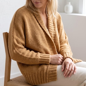 May include: A mustard yellow knit cardigan featuring a shawl collar, long sleeves with ribbed cuffs, and a ribbed hem. The garment is constructed with a stockinette stitch pattern throughout the body and sleeves, showcasing a relaxed fit.
