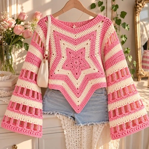 May include: A long-sleeved crochet top featuring a large central star pattern in alternating pink and cream stripes. The garment has wide, flared sleeves with an open-knit rectangular grid design and a pointed hemline, paired with light blue denim shorts.