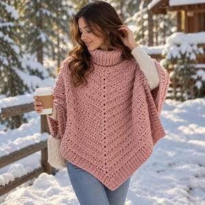 Puede incluir: Un poncho de ganchillo en color rosa empolvado con cuello alto, un patrón de puntos en forma de V texturizado y bordes acanalados. La prenda se lleva sobre una camisa blanca de manga larga y vaqueros azules, sosteniendo una taza blanca.