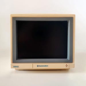 May include: A vintage Commodore 1084S color monitor featuring a beige plastic casing with a dark gray screen bezel. The front panel displays the model number 1084S on the left, the Commodore logo in the center, and a power button on the right side.