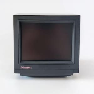 May include: A dark gray Commodore 1084S computer monitor featuring a square screen and a thick bezel. The front panel displays the Commodore logo and the model number 1084S in white text on a small rectangular label located at the bottom left corner.