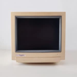 May include: A beige Commodore 1084S computer monitor with a dark gray screen bezel. The front panel features a small rectangular label in the bottom left corner with the text Commodore 1084S printed in black on a white background.