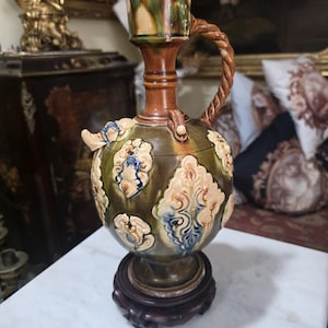 May include: A glazed ceramic ewer featuring a bulbous body with raised, cream-colored relief patterns accented in blue and green. The vessel has a tall, slender neck, a twisted rope-style handle, and a small spout, all resting on a dark wooden base.