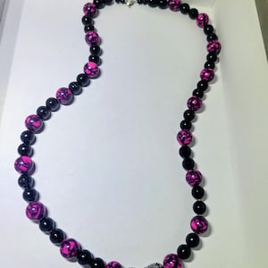 May include: A beaded necklace featuring alternating solid black beads and bright pink beads with a black crackle pattern. The necklace includes a central charm shaped like a black billiard eight ball, flanked by two small, speckled silver-toned beads.