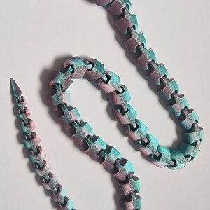 May include: An articulated snake figurine featuring a gradient colour pattern that shifts between teal and magenta. The toy is constructed from multiple interlocking segments that allow for flexible movement, ending in a pointed tail and a head.