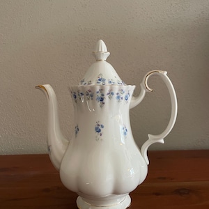 May include: A white porcelain teapot featuring a fluted body, a matching lid with a pointed finial, and a curved handle. The teapot is decorated with small blue floral patterns and delicate gold trim along the rim, base, and handle edges.