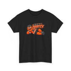 Op de afbeelding: Een zwart t-shirt met korte mouwen en een afbeelding van een harig oranje wezen met een zwarte ijshockeyhelm en zwarte broek. Het wezen ligt neer en de tekst "STAY GRITTY" staat in oranje en wit boven het personage gedrukt.