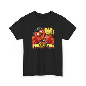 Könnte beinhalten: Ein schwarzes T-Shirt mit einer Grafik einer flauschigen orangefarbenen Kreatur, die einen schwarzen Eishockeyhelm und ein orangefarbenes Trikot mit schwarzem Logo trägt. Der Text BAD THINGS HAPPEN IN PHILADELPHIA ist gelb mit rotem Tropfeffekt.