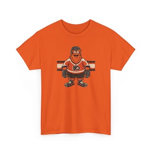 Könnte beinhalten: Ein leuchtend orangefarbenes T-Shirt mit kurzen Ärmeln und einer zentralen Grafik eines flauschigen orangefarbenen Eishockey-Maskottchens, das ein weiß-orangefarbenes Trikot mit schwarzem Logo, schwarze Shorts und einen schwarzen Helm trägt.