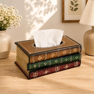 May include: A rectangular tissue box cover designed to resemble a stack of three vintage books. The top book is brown, the middle is green, and the bottom is red. Each book spine features gold-colored decorative patterns and a textured finish.