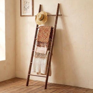 May include: A tall, dark brown wooden ladder leaning against a wall. A straw hat hangs from the top rung. A patterned orange and brown textile with fringe and a textured cream-coloured blanket are draped over the lower rungs of the ladder.
