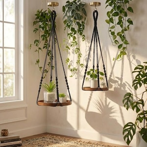 May include: Two circular wooden hanging shelves suspended by black macrame cords with knotted details. Each shelf features a gold-toned ceiling mount and holds small potted plants, including a succulent and a leafy green plant.
