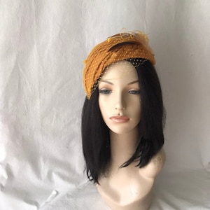 Pode incluir: Uma faixa de cabeça de feltro amarelo mostarda com um véu pequeno e penas. A faixa de cabeça é usada em um manequim com cabelo preto.