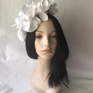 May include: White orchid flower headband for women. The headband is made of white fabric flowers and is worn on a mannequin with black hair.