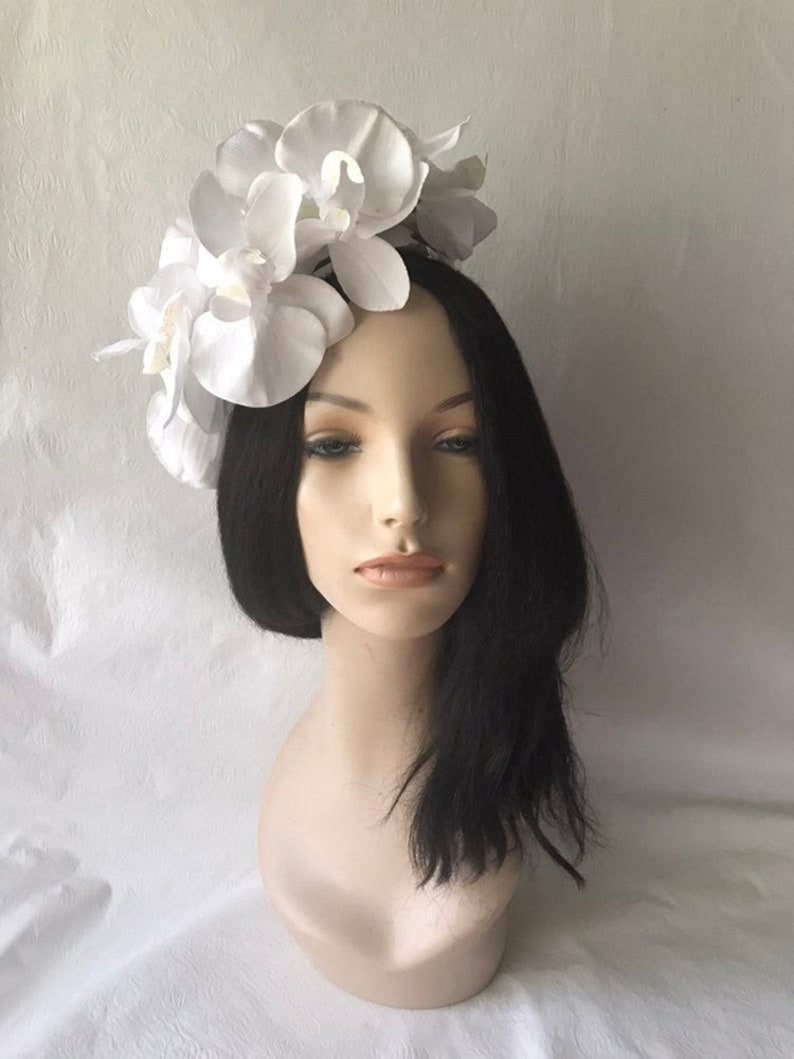 May include: White orchid flower headband for women. The headband is made of white fabric flowers and is worn on a mannequin with black hair.