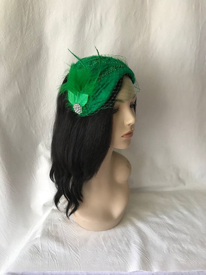 May include: A green felt hat with a green feather and a sparkly brooch. The hat is on a mannequin with long black hair.