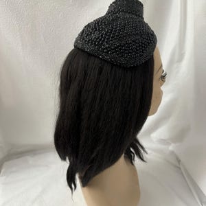 May include: A black beaded hat with a rounded, dome-like shape. The hat is made of small, black beads that are sewn together to create a textured surface.