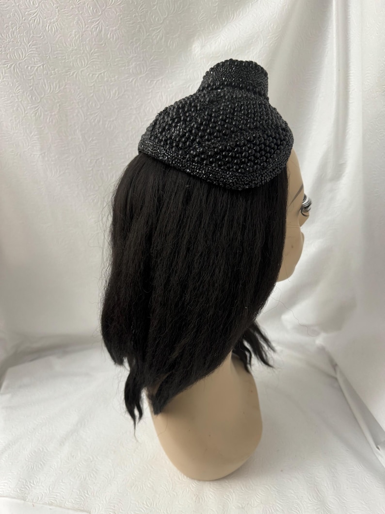 May include: A black beaded hat with a rounded, dome-like shape. The hat is made of small, black beads that are sewn together to create a textured surface.
