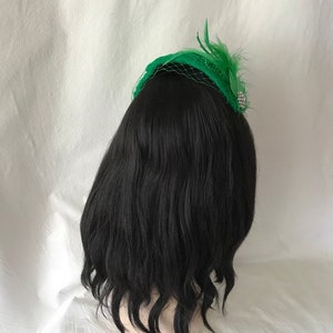 May include: A black wig with a green feather and net hat. The hat has a silver bead embellishment.
