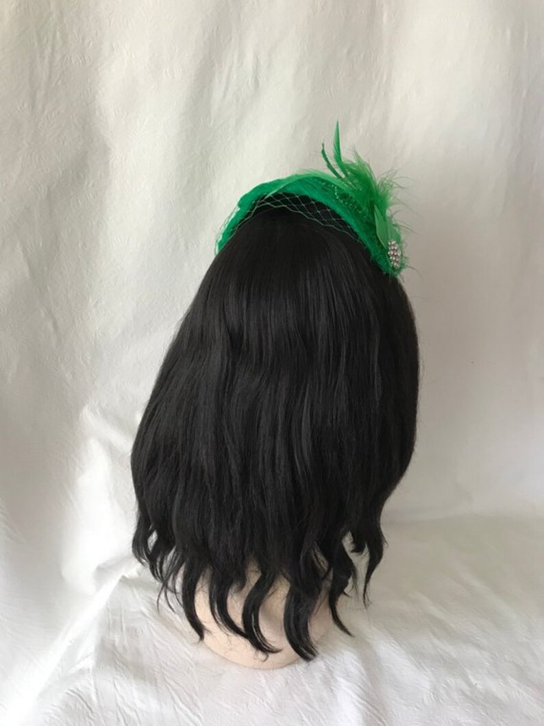 May include: A black wig with a green feather and net hat. The hat has a silver bead embellishment.