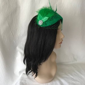 May include: A green hat with a feather and a rhinestone embellishment. The hat is made of a soft, textured fabric and has a veil.
